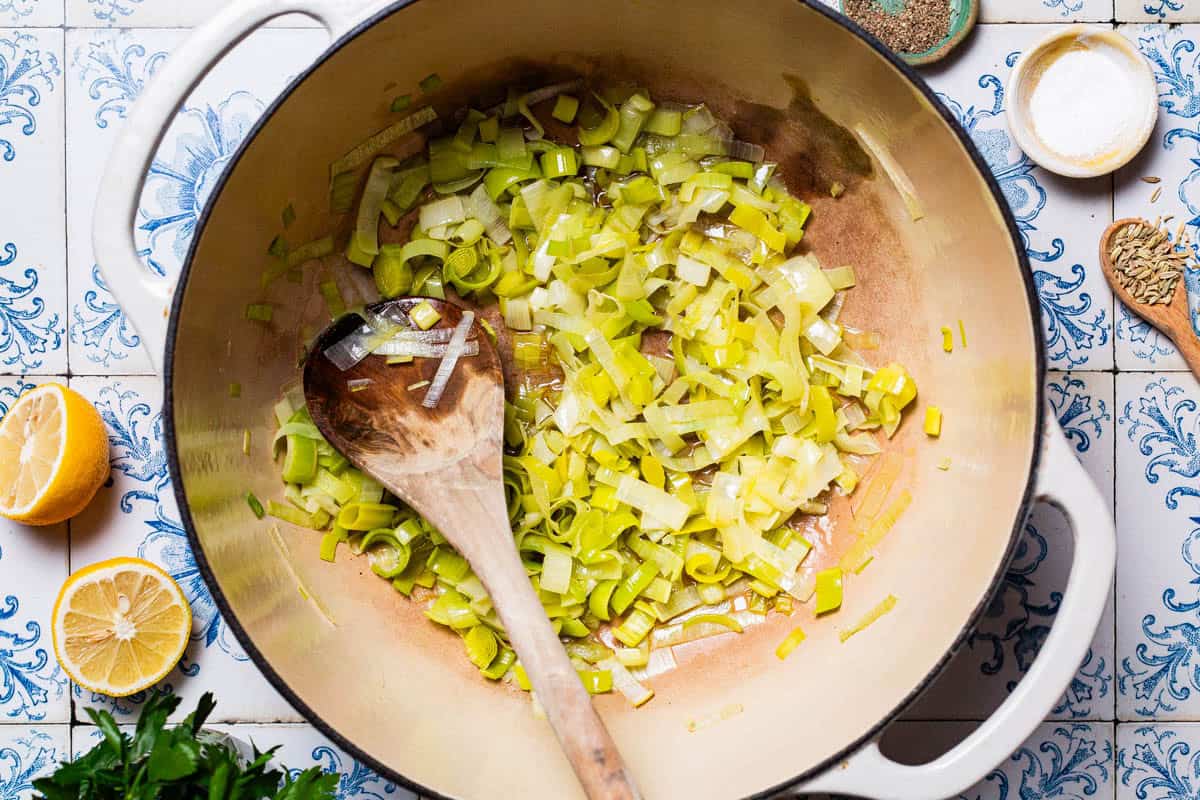 An overhead photo of chopped leeks being sauteed in a large pot with a wooden spoon. Next to this is a spoon of fennel seeds, bowls of salt and black pepper, 2 lemon halves, and some parsley.