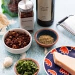 Ingredients for bread dipping oil including olive oil, sun dried tomatoes, Aleppo flakes, garlic, Italian seasoning, parsley, and parmesan cheese with a cheese grater.