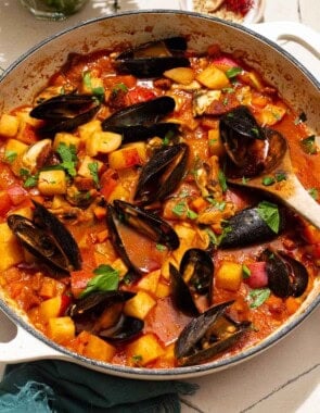 A close up of a pot mussels with chorizo in a saffron stew with a wooden spoon. Next to this a cloth napkin, bowls of spices, and some parsley.