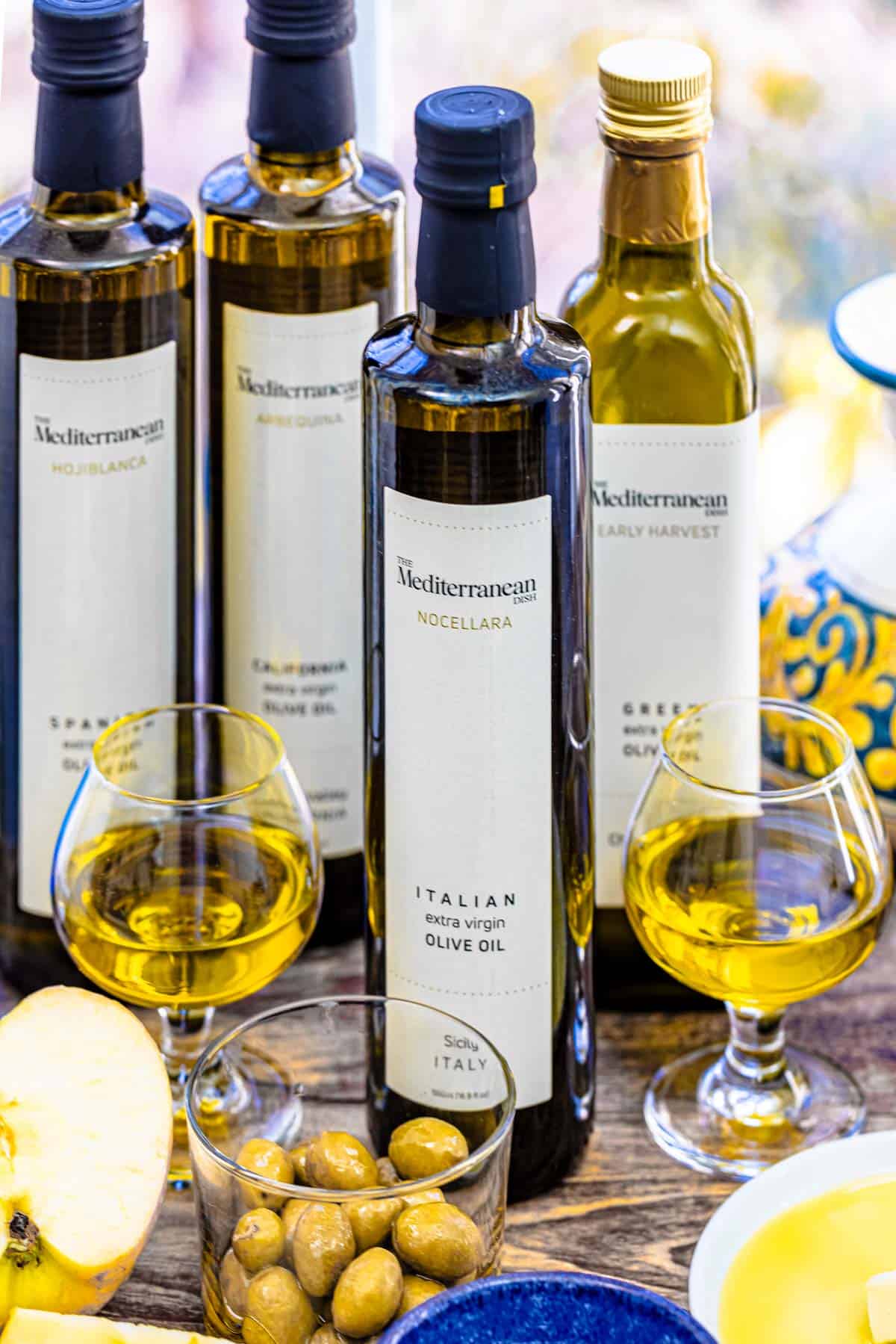4 different types of olive oils in their bottles sitting behind a half of an apple, a bowl of green olives, and two small glasses containing olive oil.