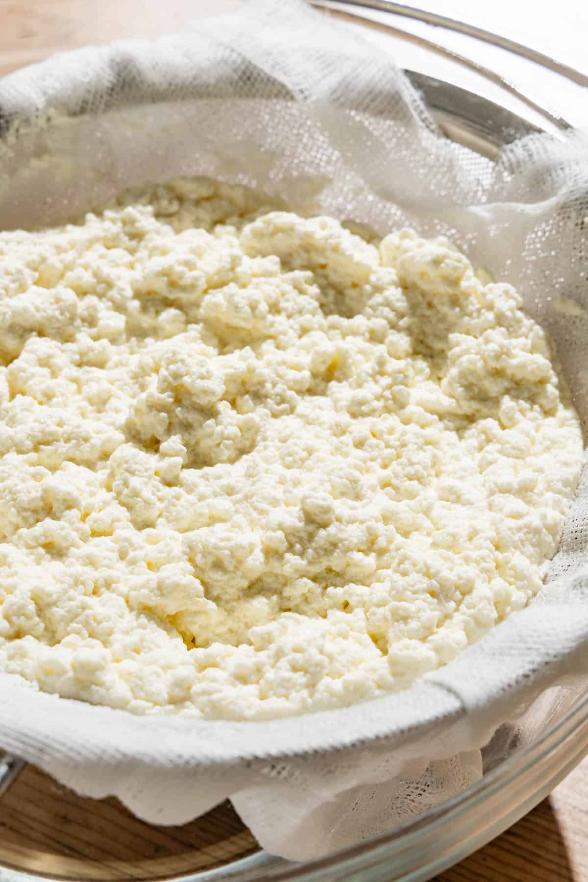 A close up of homemade ricotta being strained in a cheesecloth-lined mesh strainer sitting over a bowl.