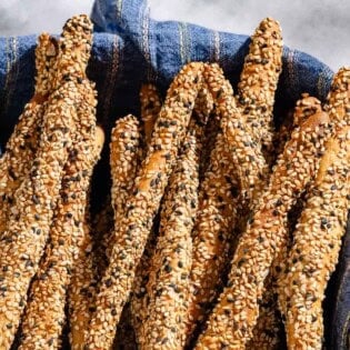 A close up of baked crispy sesame breadsticks in a basket lined with a cloth towel.