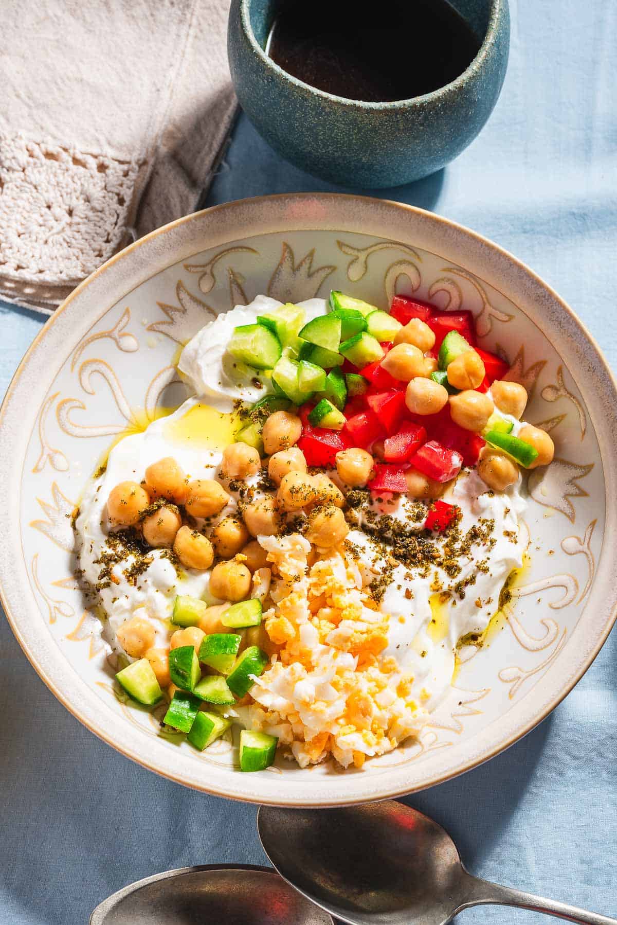 An overhead photo of a savory yogurt bowl. Next to this is a spoon, a cloth napkin, and a cup of coffee.