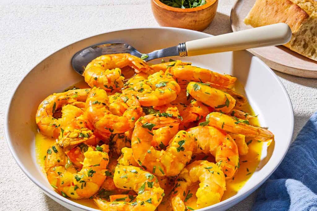 A close up of poached shrimp in a saffron and white wine sauce in a bowl with a spoon. Next to this is a bowl of chopped parsley, a cloth napkin, and some crusty bread on a plate.