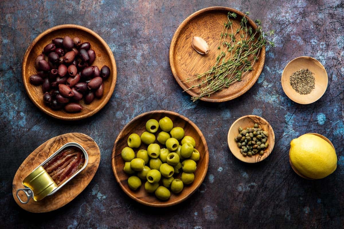 ingredients for olive tapenade including kalamata olive, castelvetrano olives, anchovies, garlic, capers, thyme, lemon, and black pepper.